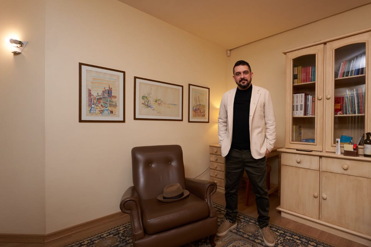 Outro ângulo do consultório, com poltrona de couro, quadros, arandela e armário de madeira com livros