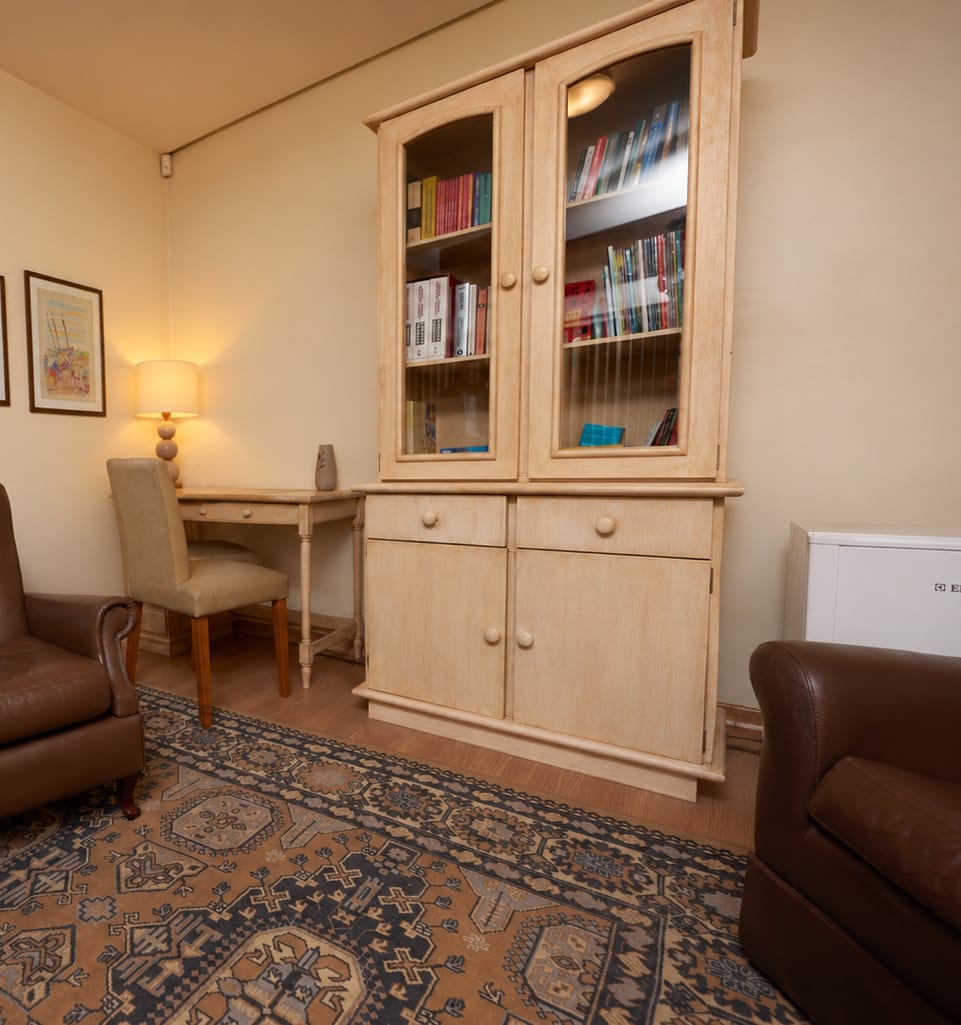 Canto da sala com mesa de apoio, cadeira, luminária acesa e armário de madeira com livros